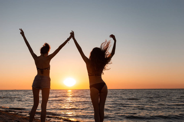 Two slim dark-haired girls in swimsuits hold hands up near the sea on the sunset - Foto, Imagem