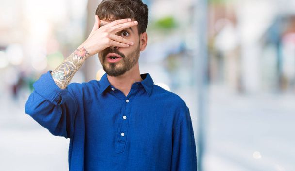 Young handsome man over isolated background peeking in shock covering face and eyes with hand, looking through fingers with embarrassed expression. - Foto, imagen