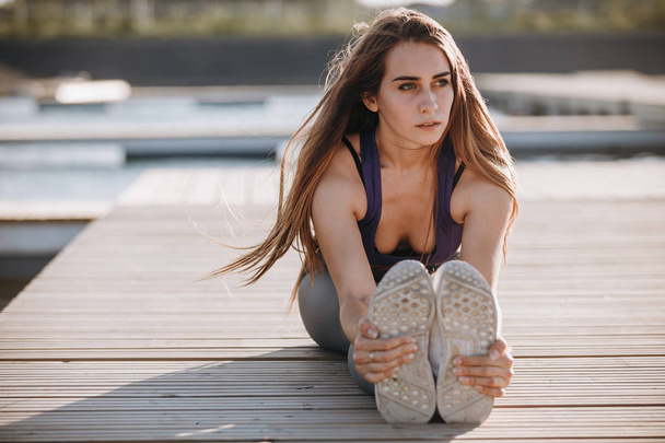 Young slim girl with long brown hair in a sports top and tights doing stretching on the wooden pier . - Foto, immagini