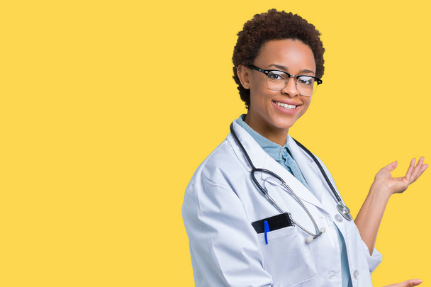 Νεαρός Αφρικανός Αμερικανός γιατρός γυναίκα, φορώντας ιατρική παλτό πάνω από απομονωμένες φόντο προσκαλεί να εισέλθουν χαμογελώντας φυσικά με ανοιχτό χέρι - Φωτογραφία, εικόνα