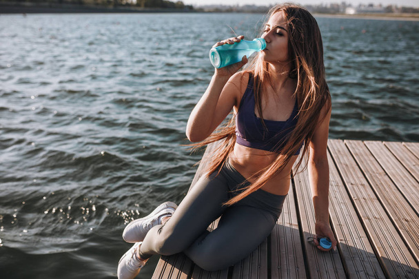 Young beautiful girl with long brown hair in a sports top and tights sits on the wooden pier and drinks water - Фото, зображення