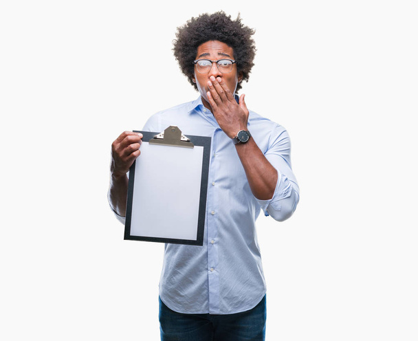 Afro american man holding clipboard over isolated background cover mouth with hand shocked with shame for mistake, expression of fear, scared in silence, secret concept - Fotografie, Obrázek