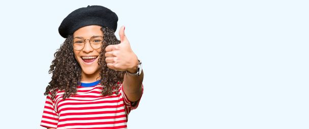 Young beautiful woman with curly hair wearing glasses and fashion beret doing happy thumbs up gesture with hand. Approving expression looking at the camera showing success. - Photo, Image