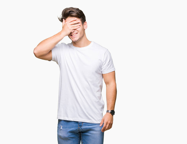Jonge knappe man dragen witte t-shirt over geïsoleerde achtergrond glimlachen en lachen met hand op gezicht die betrekking hebben op ogen voor verrassing. Blind concept. - Foto, afbeelding