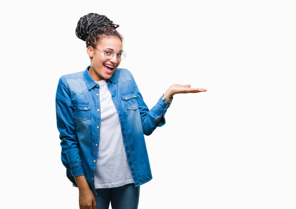 Молодий плетені волосся афро-американських дівчинка в окулярах над ізольованих фону, посміхаючись веселий подання та наведення вказівника з долоні, дивлячись на камеру. - Фото, зображення