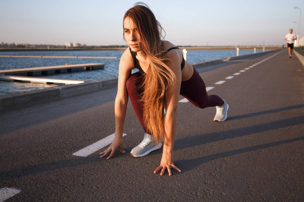 Young girl with long brown hair dressed in sports clothes doing stretching on the road along the reservoir on sunny day - Photo, image