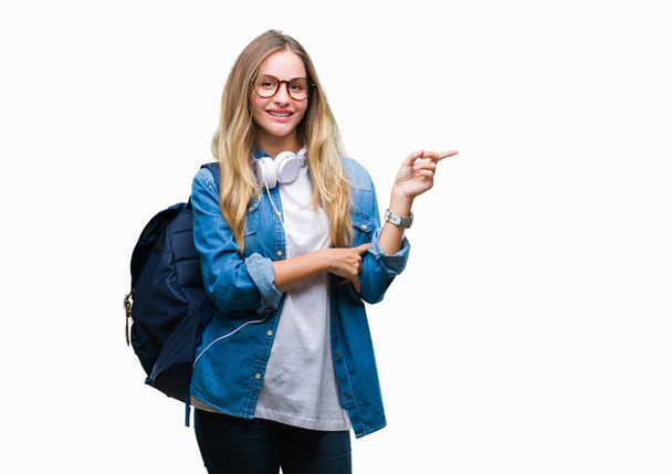 Jonge mooie blonde student vrouw hoofdtelefoons en bril dragen over geïsoleerde achtergrond met een grote glimlach op het gezicht, met de hand en de vinger te wijzen naar de kant te kijken naar de camera. - Foto, afbeelding