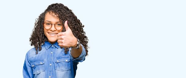 Young beautiful woman with curly hair wearing glasses doing happy thumbs up gesture with hand. Approving expression looking at the camera with showing success. - Fotografie, Obrázek
