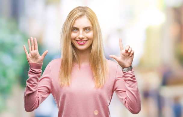 Jovem mulher loira bonita vestindo suéter de inverno rosa sobre fundo isolado mostrando e apontando para cima com os dedos número sete enquanto sorri confiante e feliz
. - Foto, Imagem