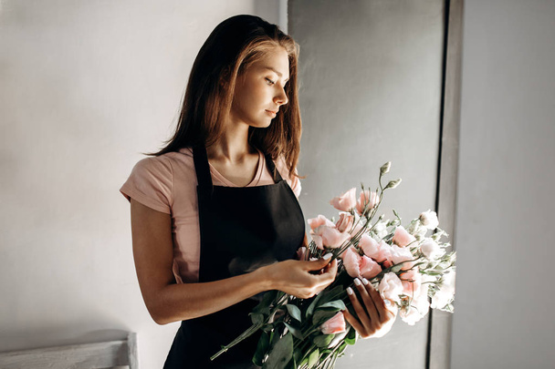 Young woman florist in a pink t-shirt and black apron holds a bouquet of eustoms in pastel colors - Valokuva, kuva
