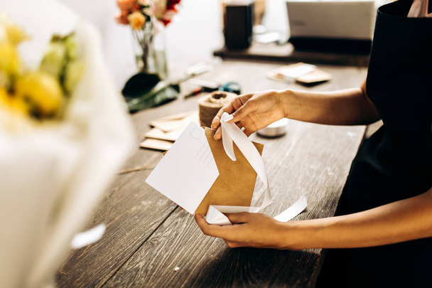 Florist is packing a postcard in an envelope with white ribbon on the wooden table - Foto, imagen