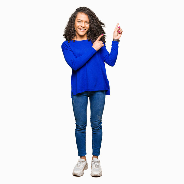 Young beautiful woman with curly hair wearing winter sweater smiling and looking at the camera pointing with two hands and fingers to the side. - Фото, изображение