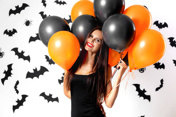 Gorgeous brunette woman in black dress holds black and orange balloons on a white background with black bats and spiders. Halloween - Photo, Image