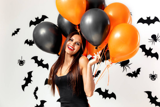 Gorgeous woman in black dress smiles and holds black and orange balloons on a white background with black bats and spiders. - Fotografie, Obrázek