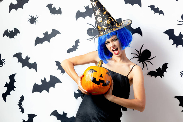 Funny young woman in a blue wig and witchs hat holds Halloween pumpkin on a white background with black bats and spiders - Zdjęcie, obraz