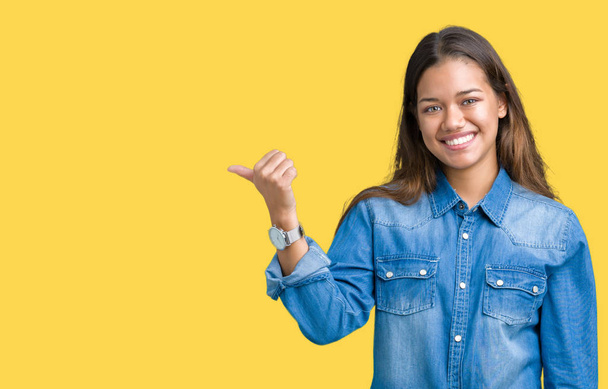 Young beautiful brunette woman wearing blue denim shirt over isolated background smiling with happy face looking and pointing to the side with thumb up. - Photo, Image