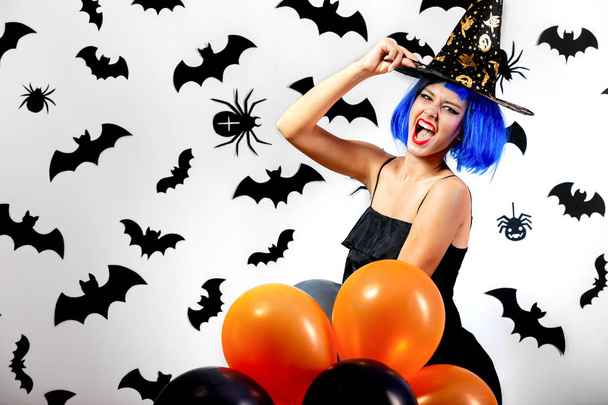 Young woman in a blue wig and witchs hat black and orange balloons on a white background with black bats and spiders - Foto, Imagen