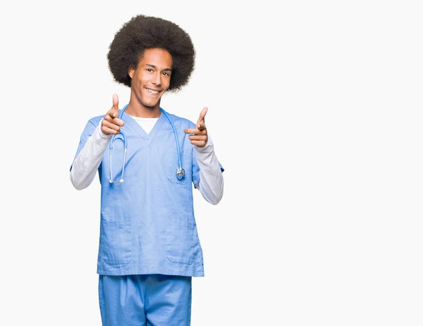 Giovane uomo medico afro americano con i capelli afro puntando il dito verso la fotocamera con viso felice e divertente. Buona energia e vibrazioni
. - Foto, immagini