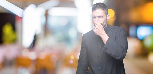 Young Christian priest over isolated background smelling something stinky and disgusting, intolerable smell, holding breath with fingers on nose. Bad smells concept. - 写真・画像