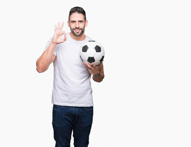 Młody człowiek posiadający piłka nożna Piłka nożna na białym tle robi ok znak palcami, doskonały symbol - Zdjęcie, obraz