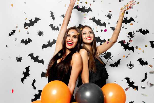 Two charming women in witches hats have fun with confetti and hold black and orange balloons on a white background with black bats and spiders. - 写真・画像