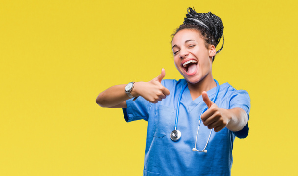 Genç örgülü saç Afro-Amerikan kız profesyonel cerrah el ile pozitif jest yapıyor onaylama izole arka plan üzerinde başparmak gülümseyen ve başarı için mutlu. Kamera, kazanan jest arıyorsunuz. - Fotoğraf, Görsel