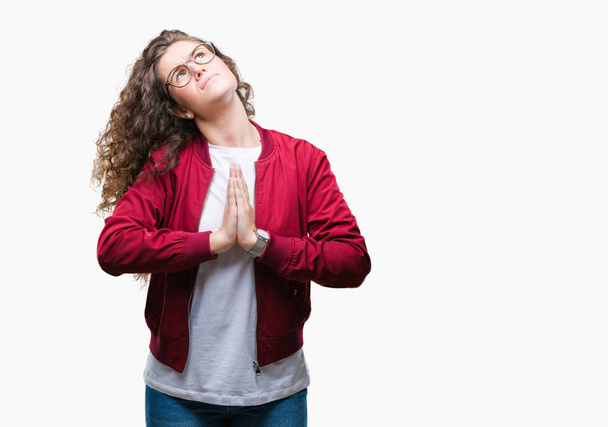 Beautiful brunette curly hair young girl wearing jacket and glasses over isolated background begging and praying with hands together with hope expression on face very emotional and worried. Asking for forgiveness. Religion concept. - Foto, immagini