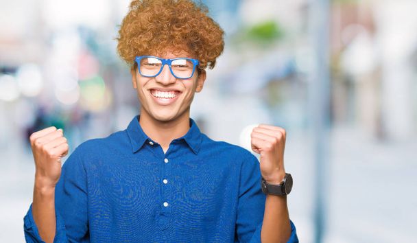 Mladý pohledný muž s afro vlasy modré brýle slaví překvapen a ohromen pro úspěch s zvedl ruce a otevřít oči. Vítěz koncept. - Fotografie, Obrázek