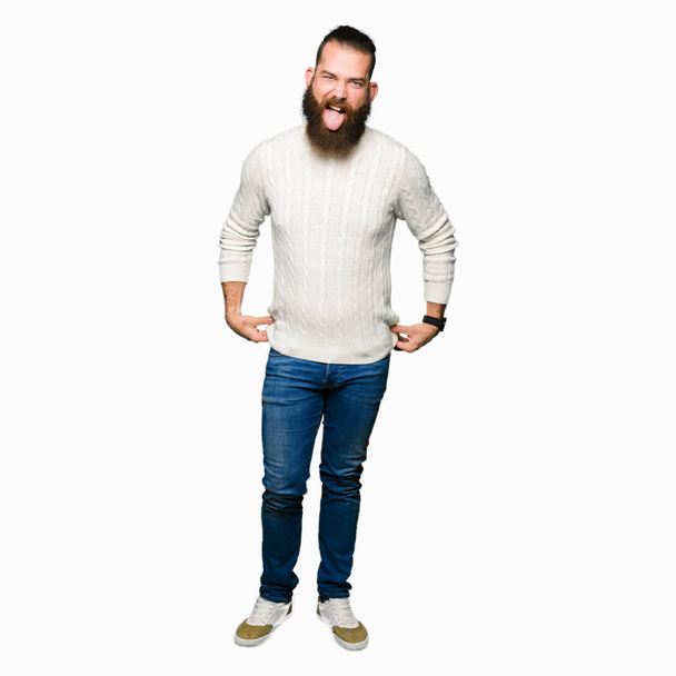 Jonge hipster man dragen winter trui steken tong uit blij met grappige expressie. Emotie concept. - Foto, afbeelding