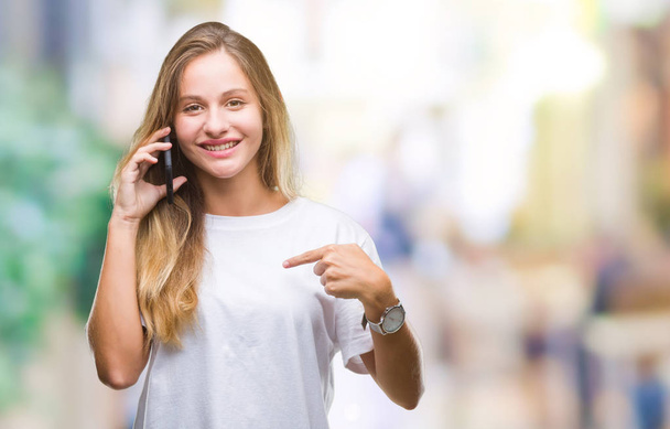 Young beautiful blonde woman calling using smartphone over isolated background with surprise face pointing finger to himself - Photo, Image