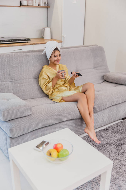 Happy woman with a remote control, choosing a TV channel during a rest after a bath. On the head is a towel. - Foto, Imagen