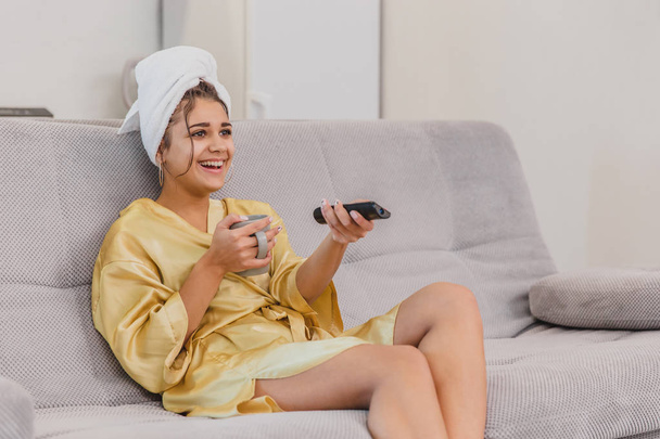 Happy woman with a remote control, choosing a TV channel during a rest after a bath. On the head is a towel. - Fotó, kép