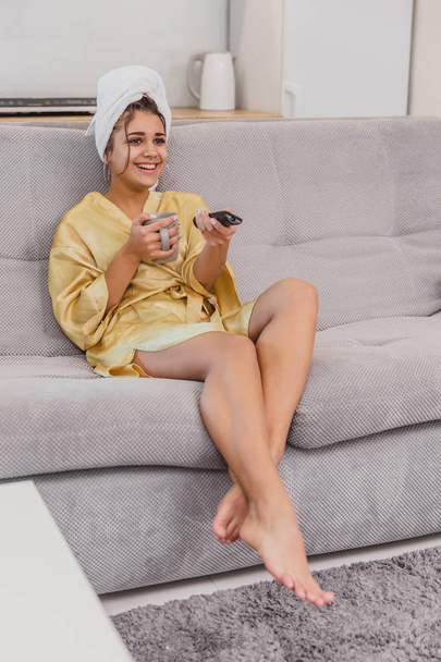 Happy woman with a remote control, choosing a TV channel during a rest after a bath. On the head is a towel. - Φωτογραφία, εικόνα