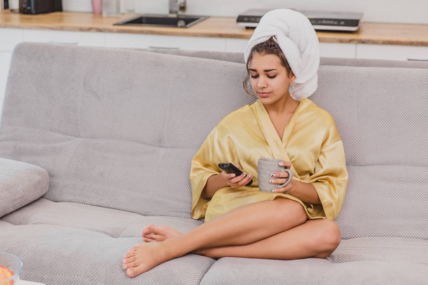 A happy woman with a remote control, choosing a television channel, holds the cup in her hand. During the rest after the bath. - Fotografie, Obrázek
