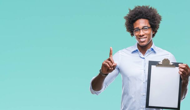 Hombre afroamericano sosteniendo portapapeles sobre fondo aislado sorprendido con una idea o pregunta apuntando con el dedo con la cara feliz, número uno
 - Foto, Imagen