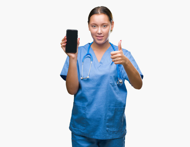 Young caucasian doctor woman showing smarpthone over isolated background happy with big smile doing ok sign, thumb up with fingers, excellent sign - Foto, imagen