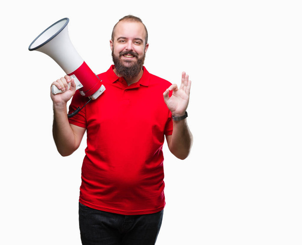 Young caucasian man shouting through megaphone over isolated background doing ok sign with fingers, excellent symbol - Photo, Image