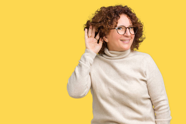 Beautiful middle ager senior woman wearing turtleneck sweater and glasses over isolated background smiling with hand over ear listening an hearing to rumor or gossip. Deafness concept. - Photo, Image