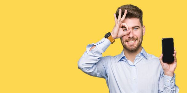 Jeune homme beau entreprise montrant écran de smartphone avec le visage heureux souriant faire ok signe avec la main sur les yeux en regardant à travers les doigts
 - Photo, image