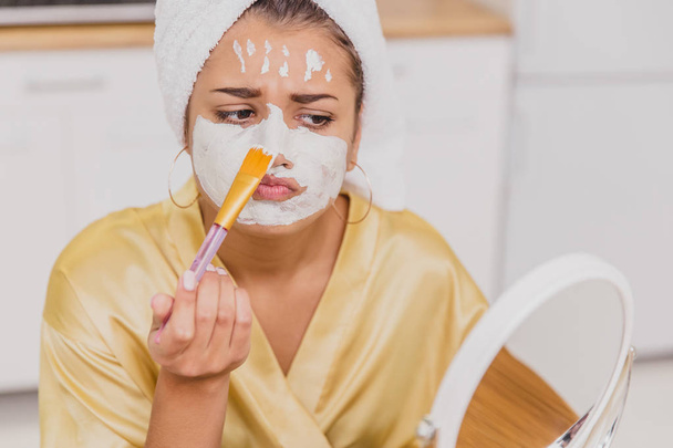Face and body skin care, face treatment at home concept. Happy young woman applying a white mask on her face with a brush. - Photo, image