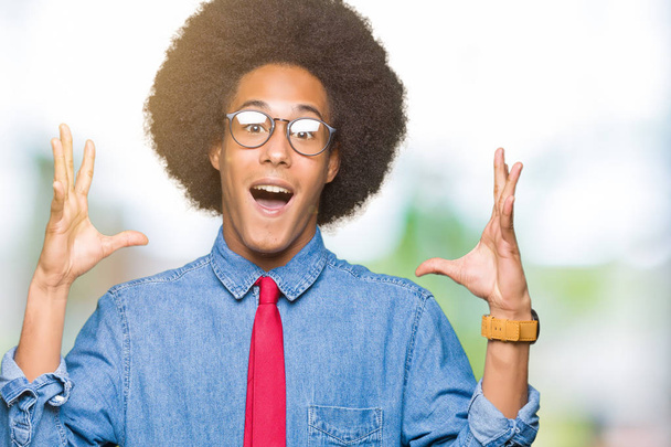 Jeune homme d'affaires afro-américain aux cheveux afro portant des lunettes et cravate rouge célébrant fou et étonné pour le succès avec les bras levés et les yeux ouverts hurlant excités. Concept gagnant
 - Photo, image