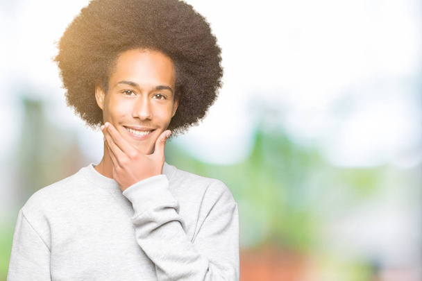 Young african american man with afro hair wearing sporty sweatshirt looking confident at the camera with smile with crossed arms and hand raised on chin. Thinking positive. - Photo, Image
