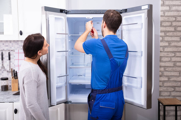 Woman Looking At Male Repairman Fixing Refrigerator With Screwdriver - 写真・画像