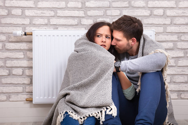 Young Couple Wrapped In Blanket Sitting In Front Of Radiator Against Brick Wall At Home - Photo, image