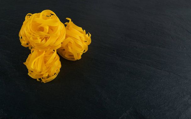 Желтый Fabrican pasta fettuccine, fettuccelle или tagliatelle крупным планом. Домашняя сухая лапша из яиц, длиннокатаные макароны или сырые спагетти на фоне черного камня
 - Фото, изображение