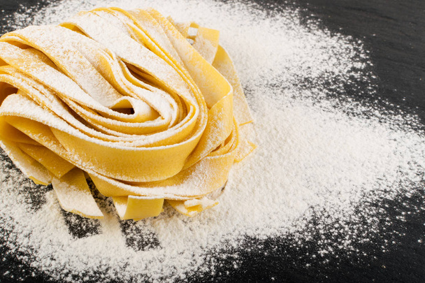 Raw yellow italian pasta pappardelle, fettuccine or tagliatelle close up. Egg homemade noodles cooking process, long rolled macaroni or uncooked spaghetti on black stone background - Foto, imagen
