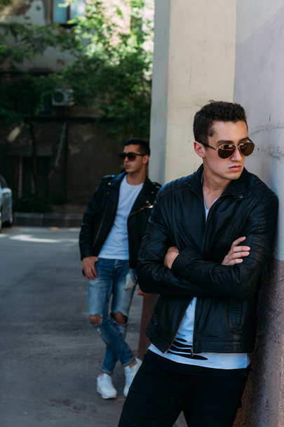 beautiful and attractive guy is standing in the city, posing. dressed in a black leather jacket, white T-shirt, jeans and white shoes. near the mirror building. walk around the city in black glasses. communication. meeting friends. - Photo, image