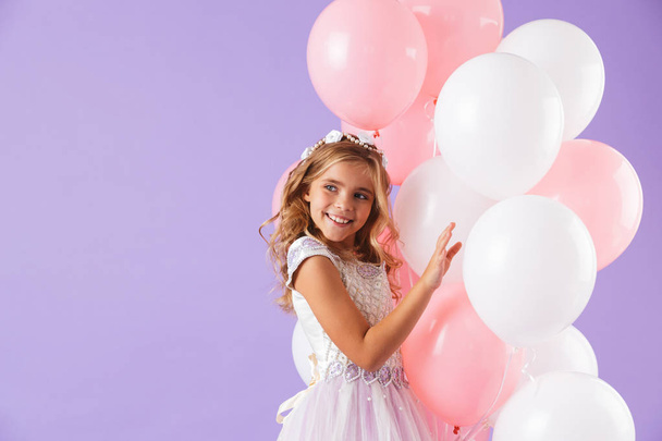 Cute pretty little girl dressed in princess dress isolated over violet background, holding bunch of balloons - Фото, изображение