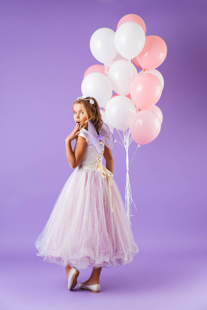 Full length of a pretty little girl dressed in princess dress standing isolated over violet background, holding bunch of balloons - Fotó, kép