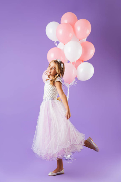Full length of a pretty little girl dressed in princess dress standing isolated over violet background, holding bunch of balloons - Фото, изображение